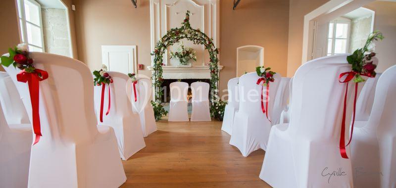 Chaises blanches décorées de rubans rouges et de fleurs, disposées face à une arche florale dans une salle lumineuse.