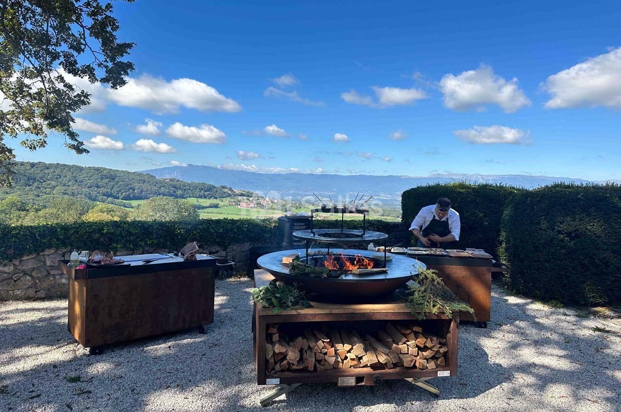 Chef cuisinant en extérieur sur des grills modernes, entouré de verdure et d'un paysage vallonné sous un ciel bleu.