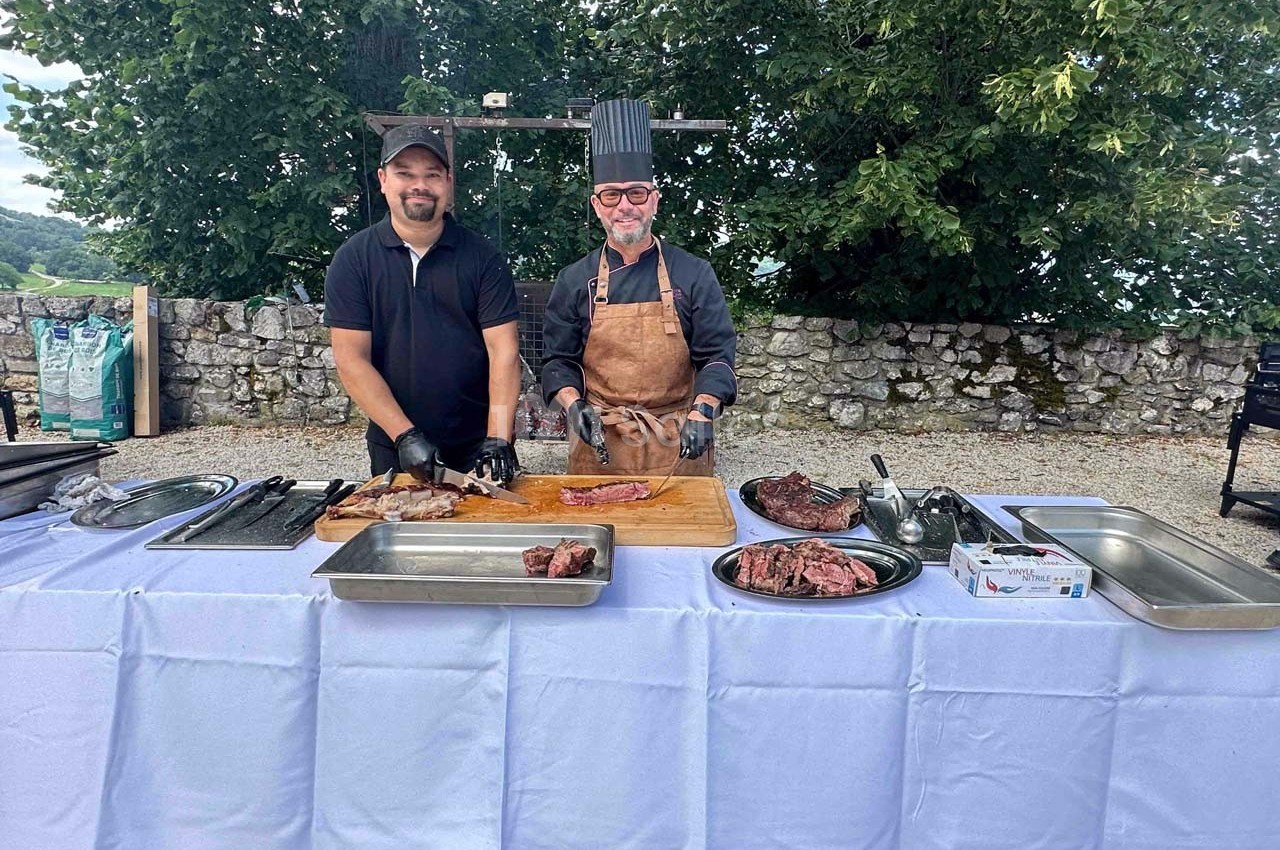 Deux hommes souriants préparent et présentent des morceaux de viande sur une table dressée en extérieur.