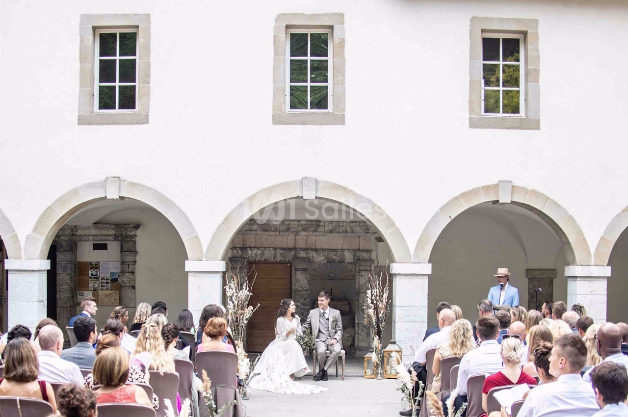 Cérémonie de mariage en extérieur sous des arches, avec les mariés assis face à des invités attentifs.