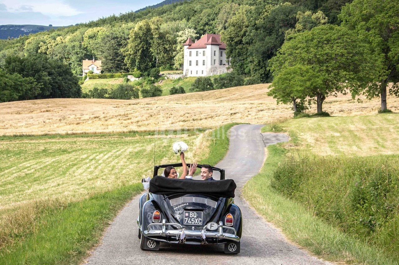 Un couple dans une voiture décapotable ancienne roule sur un chemin de campagne, avec un manoir en arrière-plan.