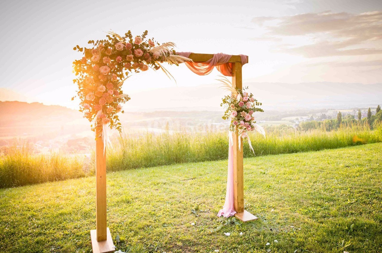 Arche en bois décorée de fleurs roses et de tissu, installée sur une pelouse au coucher du soleil.