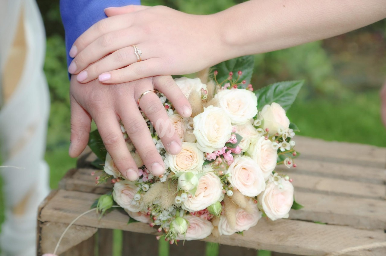 Deux mains avec des alliances posées sur un bouquet de roses blanches et roses, sur une table en bois.