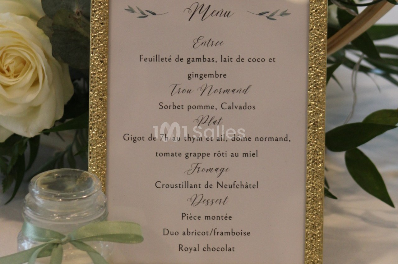 Menu de mariage encadré doré avec des fleurs blanches en arrière-plan et un pot décoratif au ruban vert.