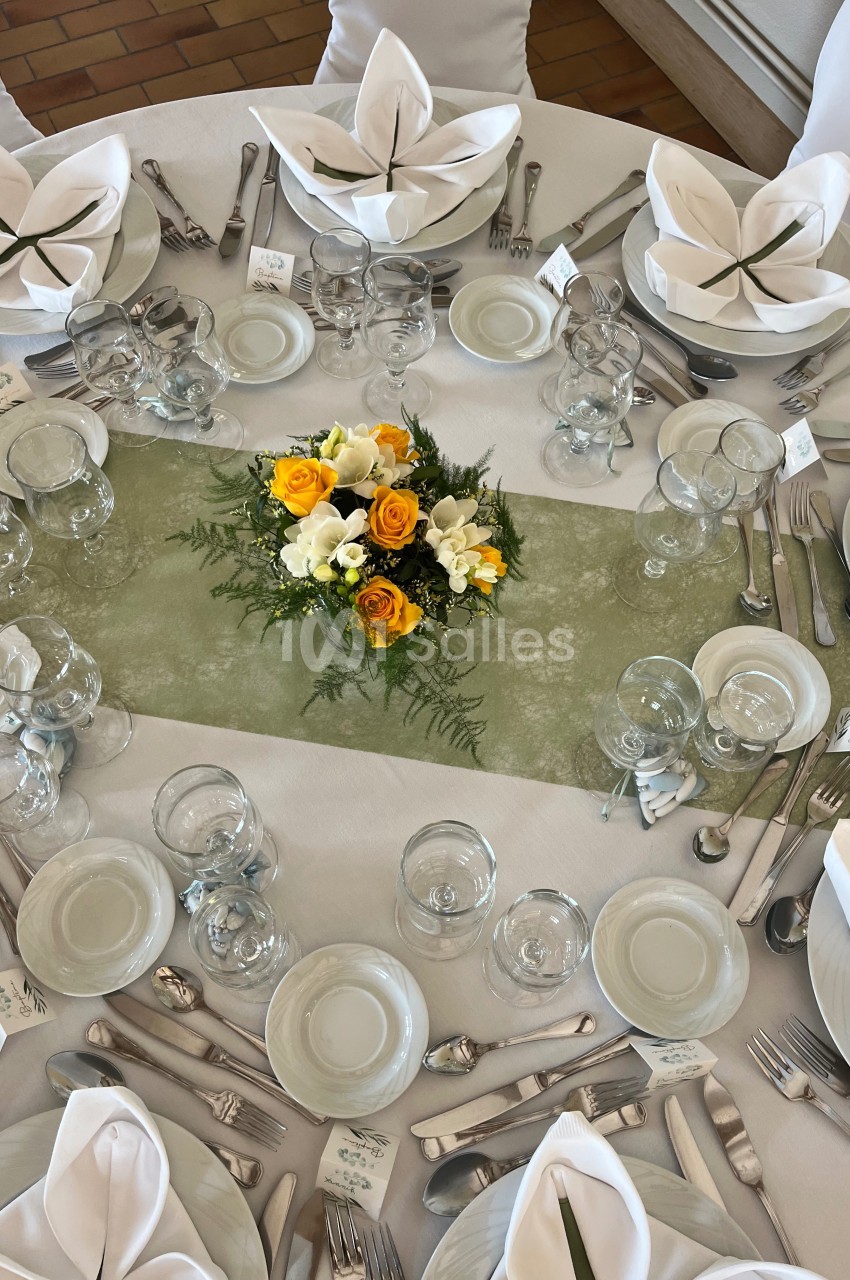 Table dressée avec nappes blanches, serviettes pliées, couverts alignés et centre de table floral jaune et blanc.