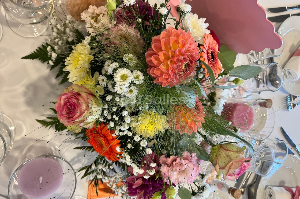 Bouquet coloré de fleurs variées, posé sur une table dressée avec verres et décorations.