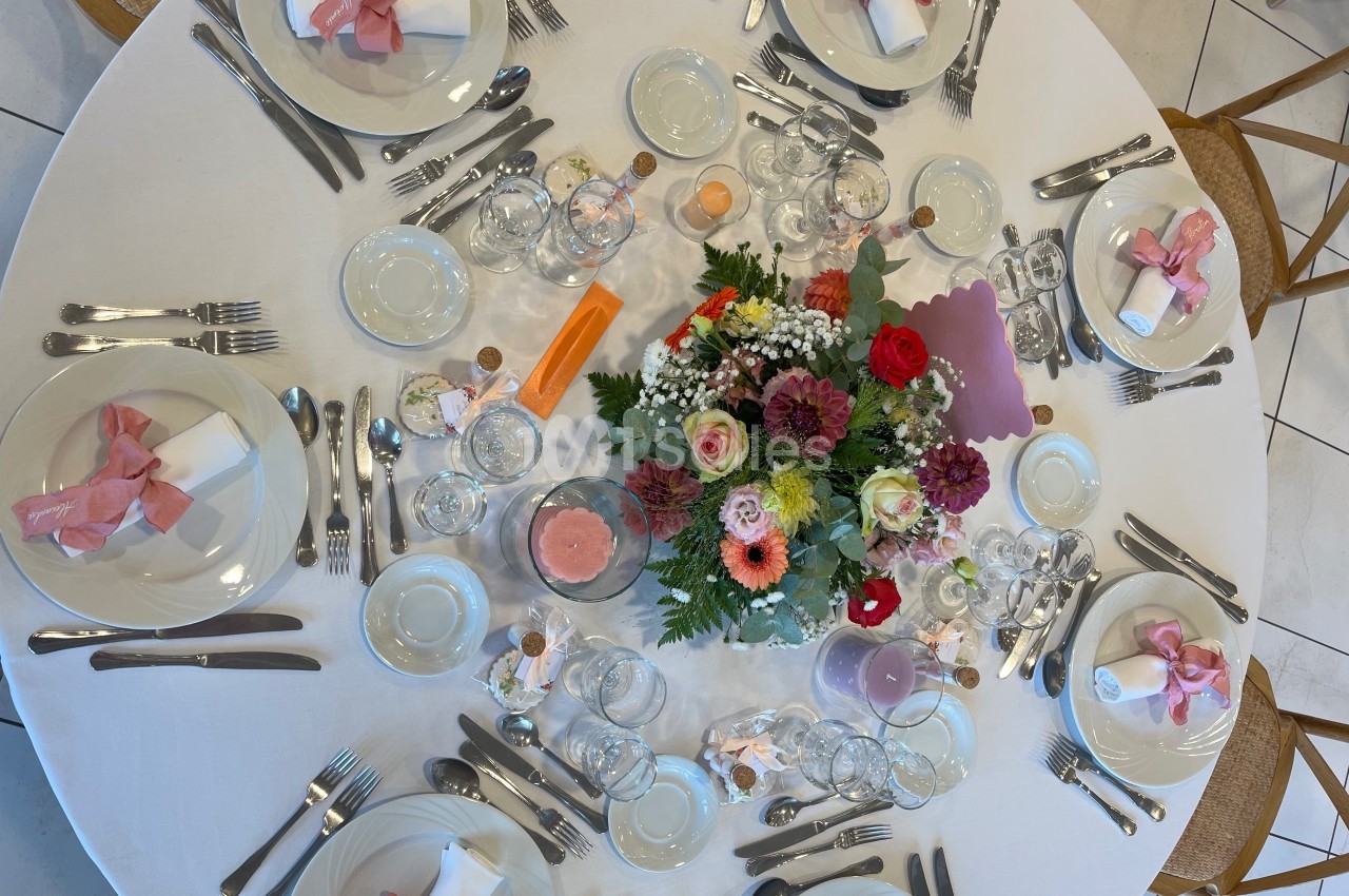 Table ronde élégamment dressée avec vaisselle, couverts, serviettes roses et centre de table floral coloré.