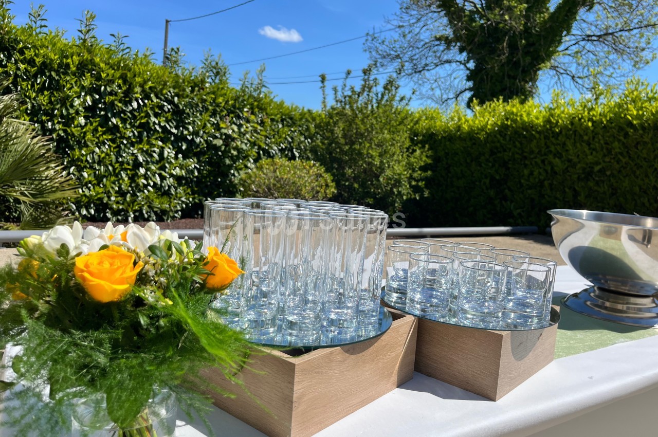 Verres empilés sur des plateaux en bois, avec un bouquet de fleurs et un bol en métal, en extérieur sous un ciel bleu.