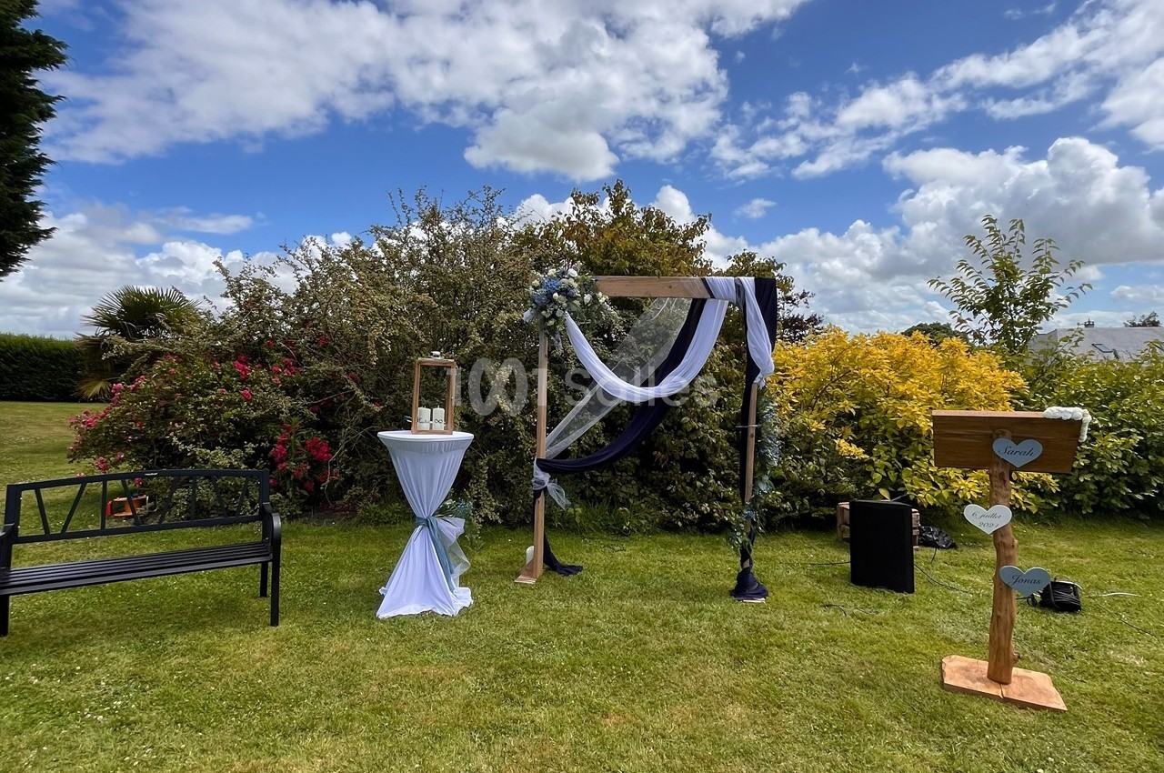 Arche de mariage décorée de tissu bleu et blanc dans un jardin fleuri sous un ciel partiellement nuageux.
