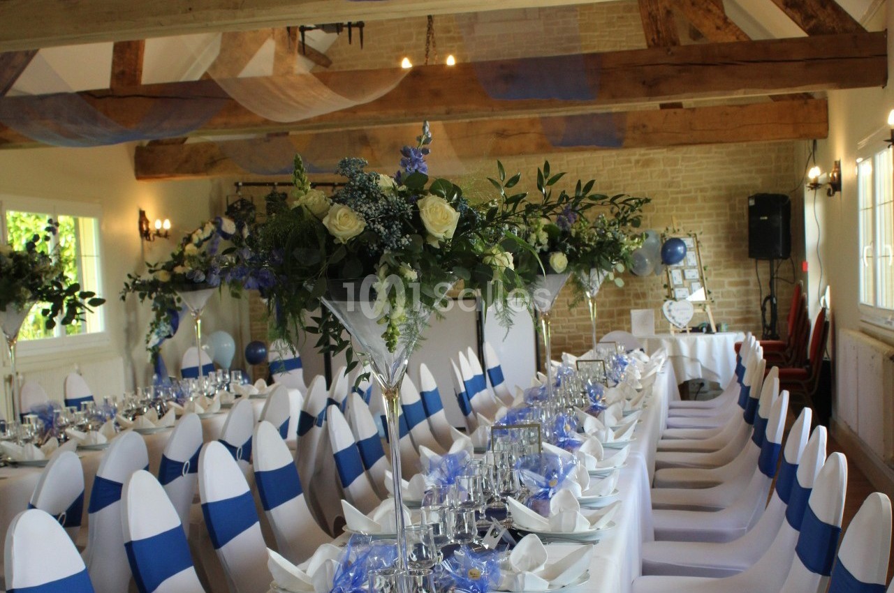 Salle de réception décorée pour un événement, avec des tables dressées, des chaises blanches ornées de rubans bleus et des…
