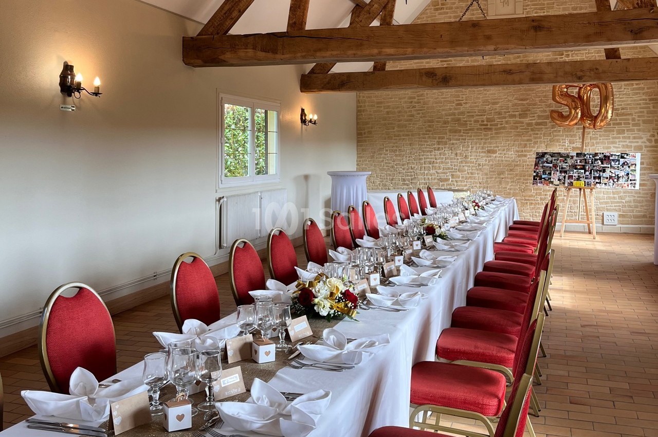 Salle décorée pour une réception avec une longue table dressée, chaises rouges et ballons dorés formant ’50’.