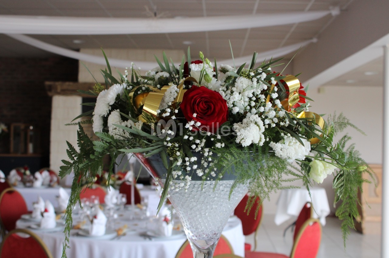 Centre de table floral avec roses rouges, fleurs blanches et rubans dorés, dans une salle de réception décorée.