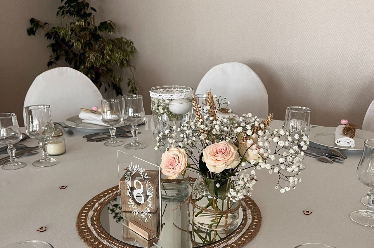 Centre de table avec roses, gypsophile, bougie et miroir, entouré de verres et assiettes sur une nappe beige.