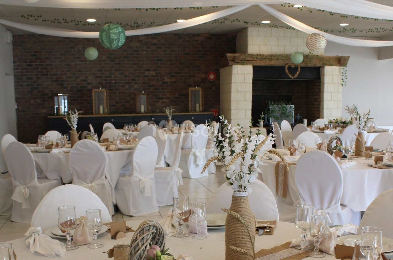 Salle de réception décorée avec des tables rondes, nappes blanches, centres de table floraux et guirlandes suspendues.