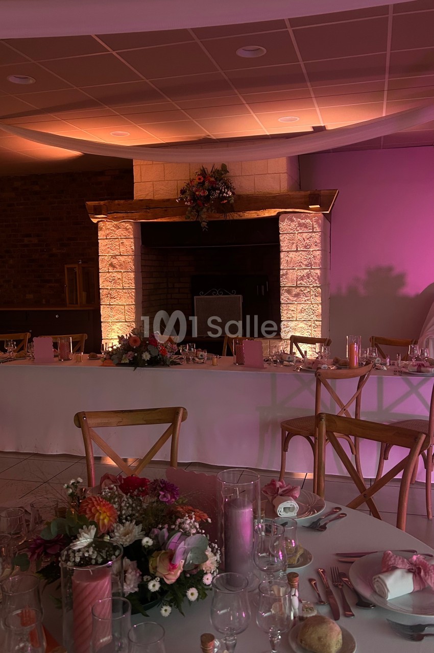 Salle de réception décorée avec des tables dressées, des fleurs et un éclairage chaleureux près d'une cheminée en pierre.
