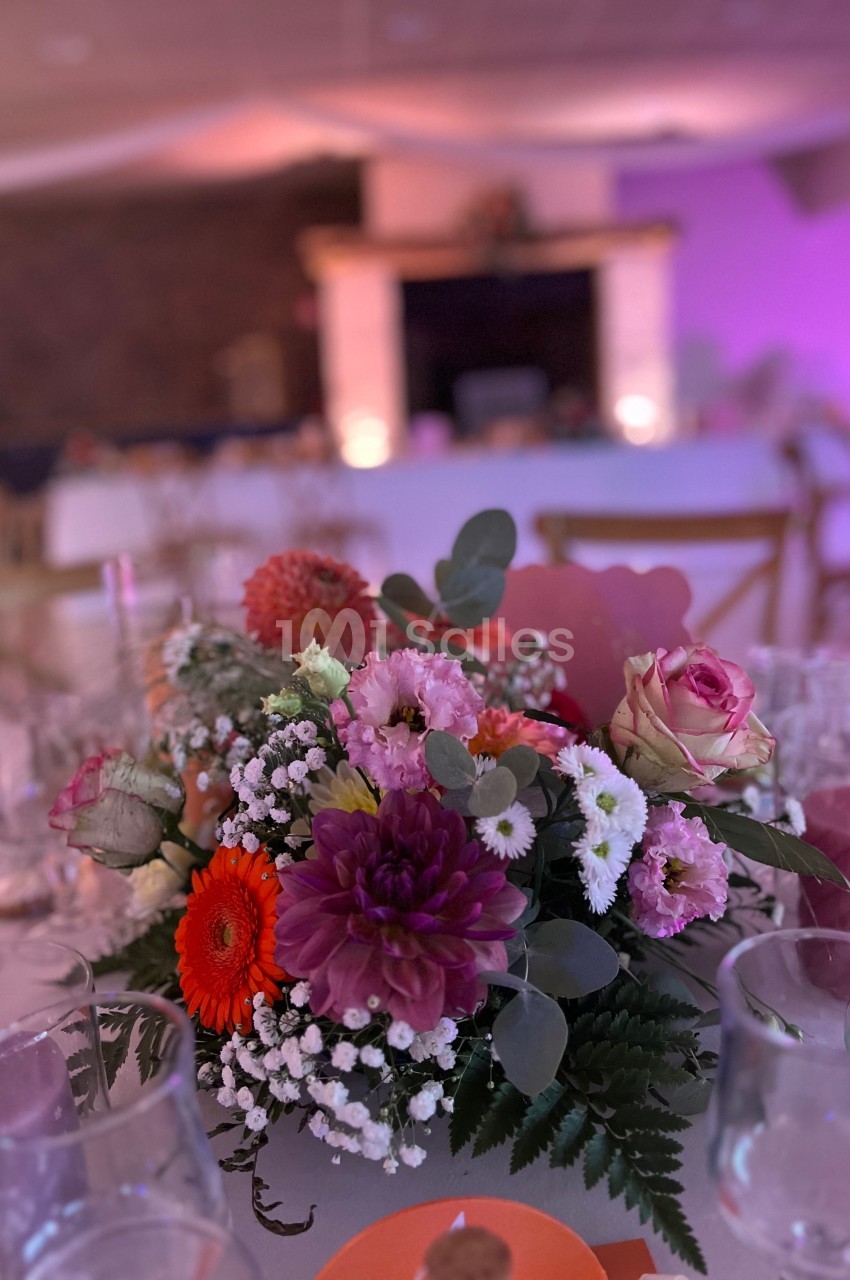Centre de table floral avec des fleurs colorées, entouré de verres et d'une ambiance lumineuse tamisée.