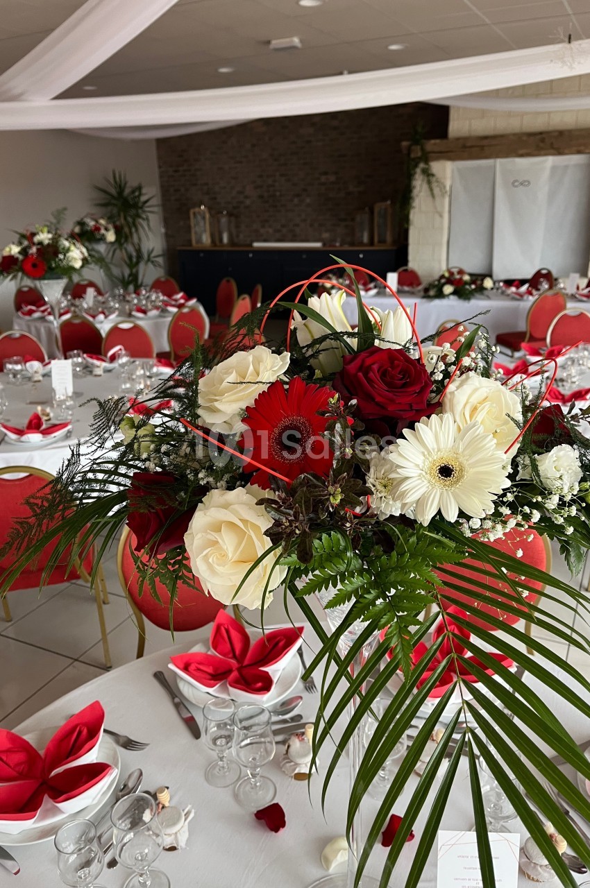 Centre de table floral avec roses rouges, gerberas blancs et feuillage, dans une salle de réception décorée.