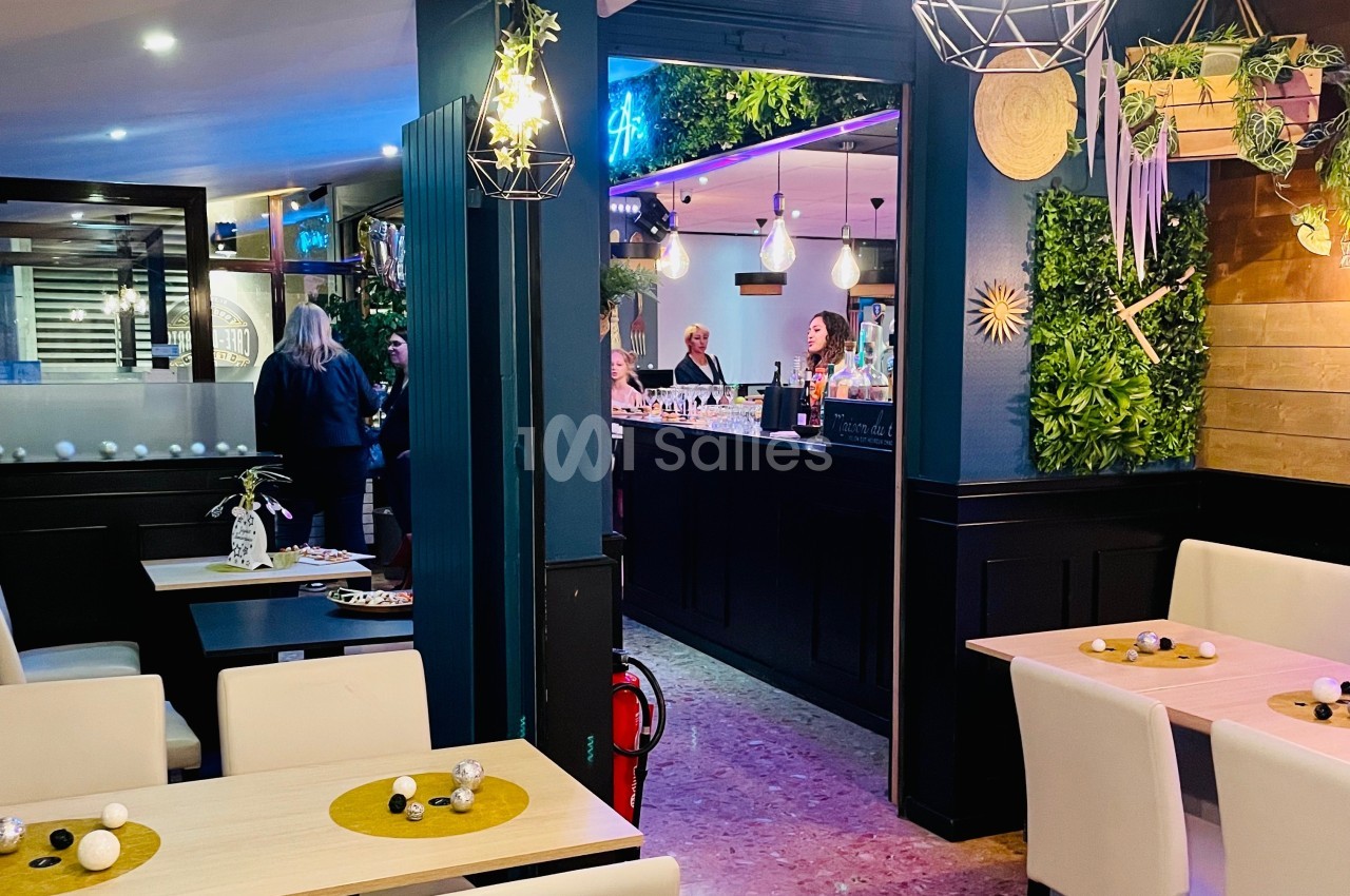 Intérieur d'un restaurant moderne avec tables dressées, décoration végétale et vue sur un bar animé en arrière-plan.