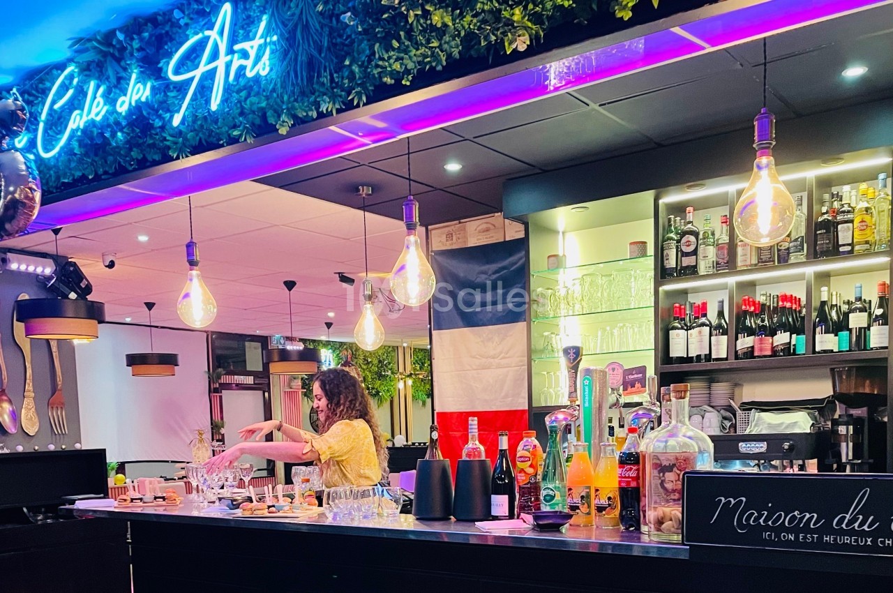 Bar lumineux avec une femme préparant des boissons, décoré de bouteilles, plantes et une enseigne néon ’Café des Arts’.