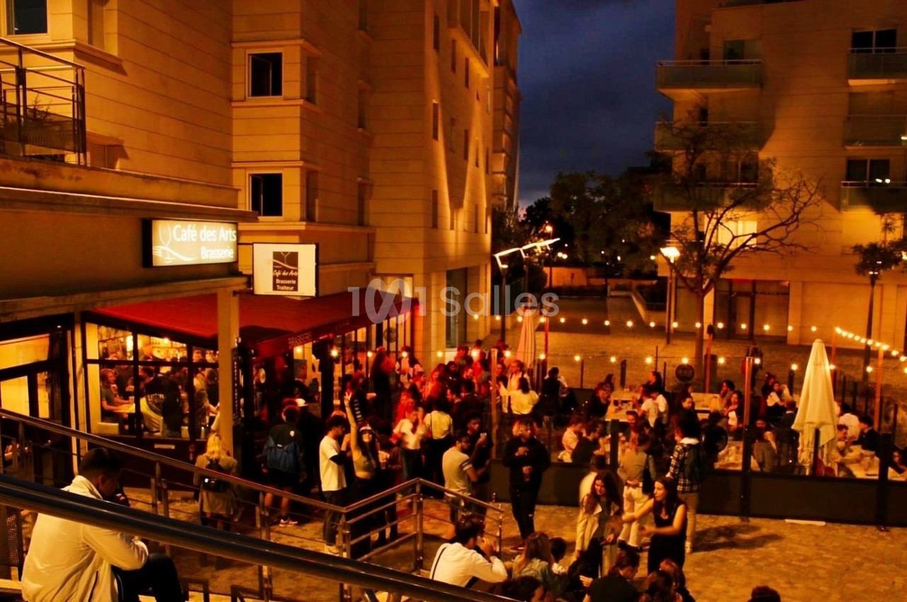 Terrasse animée d'un café en soirée, éclairée par des guirlandes lumineuses, avec de nombreuses personnes rassemblées.