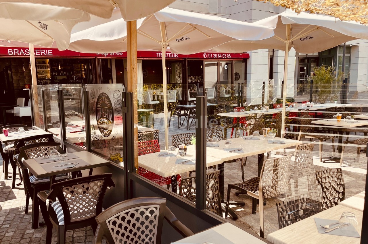 Terrasse ensoleillée d'une brasserie avec tables dressées, chaises modernes et parasols, située sur une place pavée.