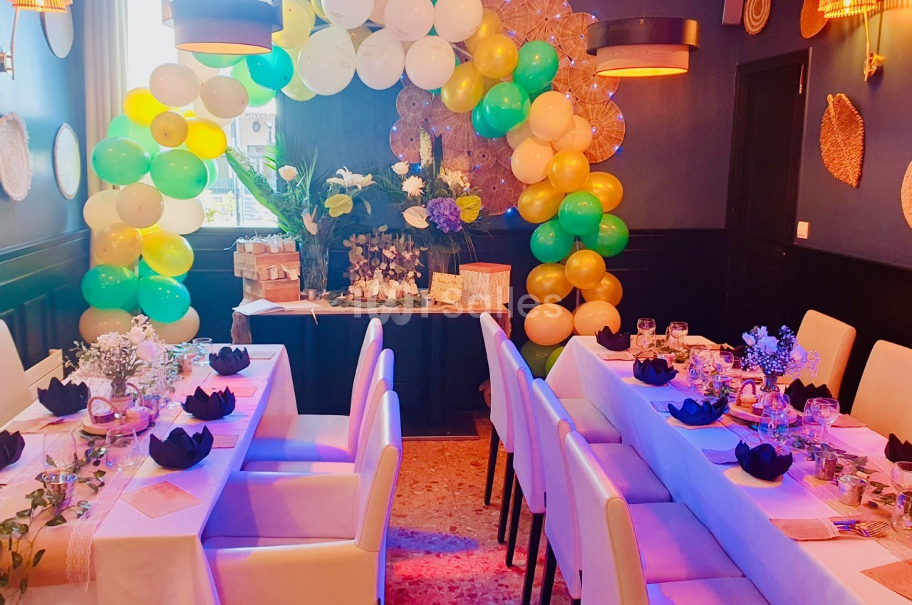 Salle décorée pour une réception avec des tables dressées, des ballons colorés et une arche florale en arrière-plan.