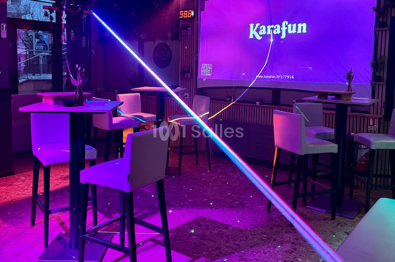 Salle de karaoké avec éclairage coloré, tables hautes, chaises blanches et écran affichant ’Karafun’.
