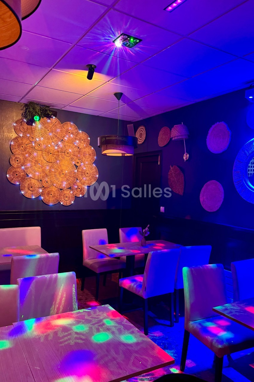 Salle de restaurant avec tables, chaises et décorations murales, éclairée par des lumières colorées.