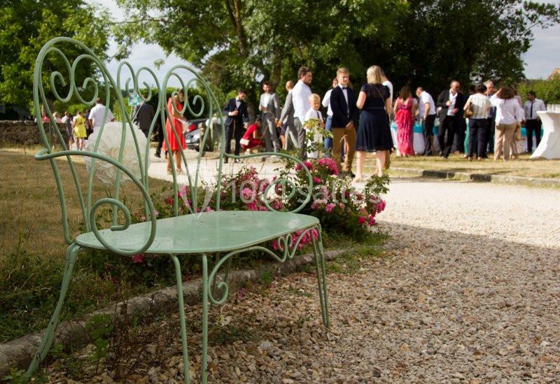 Banc en fer forgé vert clair devant des fleurs, avec un groupe de personnes rassemblées en arrière-plan dans un jardin.