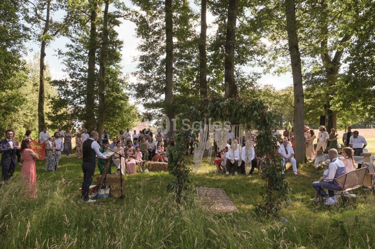 Cérémonie en plein air dans un cadre boisé, avec des invités assis et un officier célébrant devant une arche décorée.
