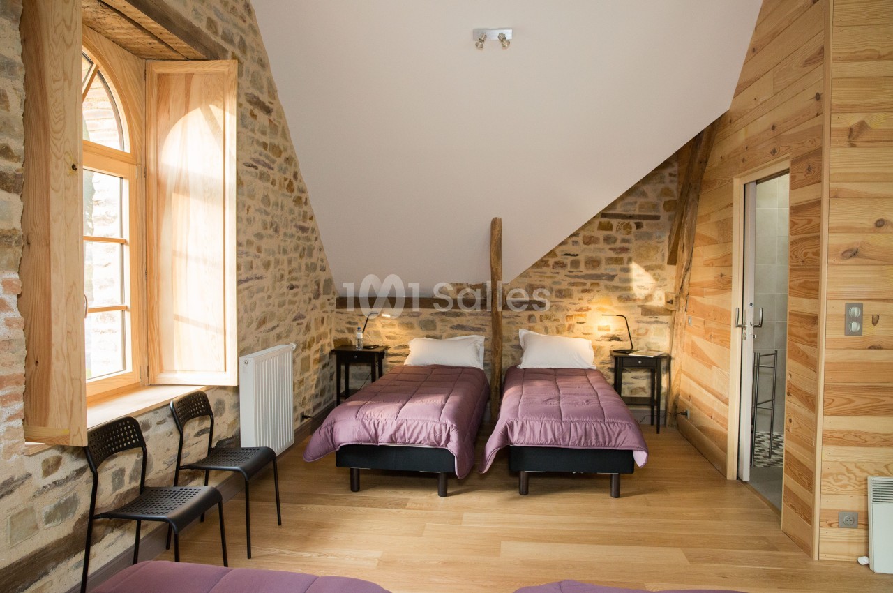 Chambre avec deux lits simples, murs en pierre et bois, fenêtre en ogive laissant entrer la lumière naturelle.
