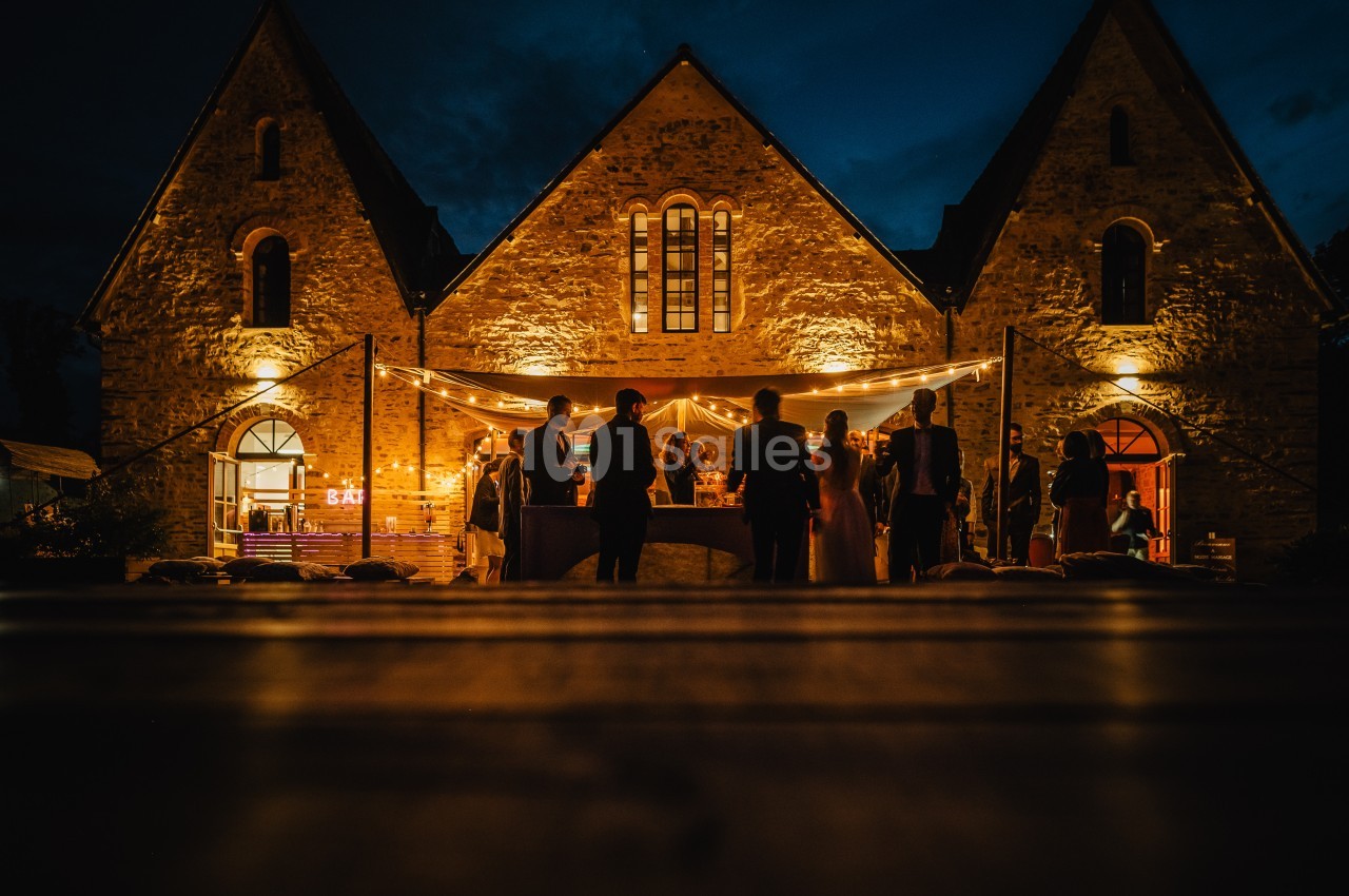 Groupe de personnes rassemblées devant un bâtiment en pierre éclairé, avec des guirlandes lumineuses, en soirée.