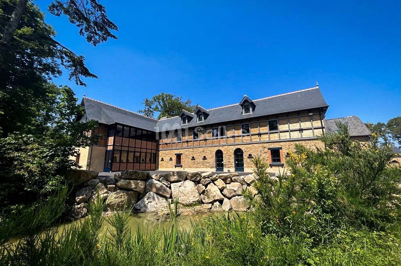 Bâtiment en briques et verre entouré de végétation, avec un ruisseau et des rochers au premier plan sous un ciel bleu.