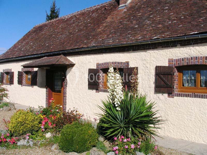 Façade d'une maison traditionnelle avec murs clairs, volets en bois, toit en tuiles et jardin fleuri au premier plan.