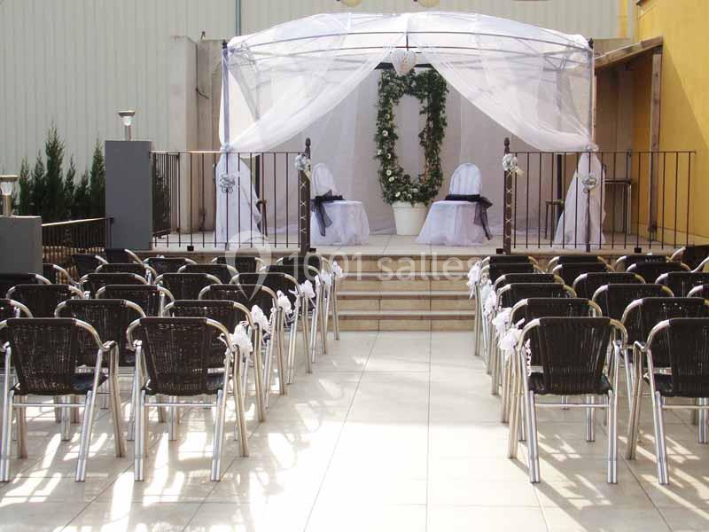 Chaises alignées devant une arche décorée de fleurs blanches sous une tonnelle, préparée pour une cérémonie en extérieur.