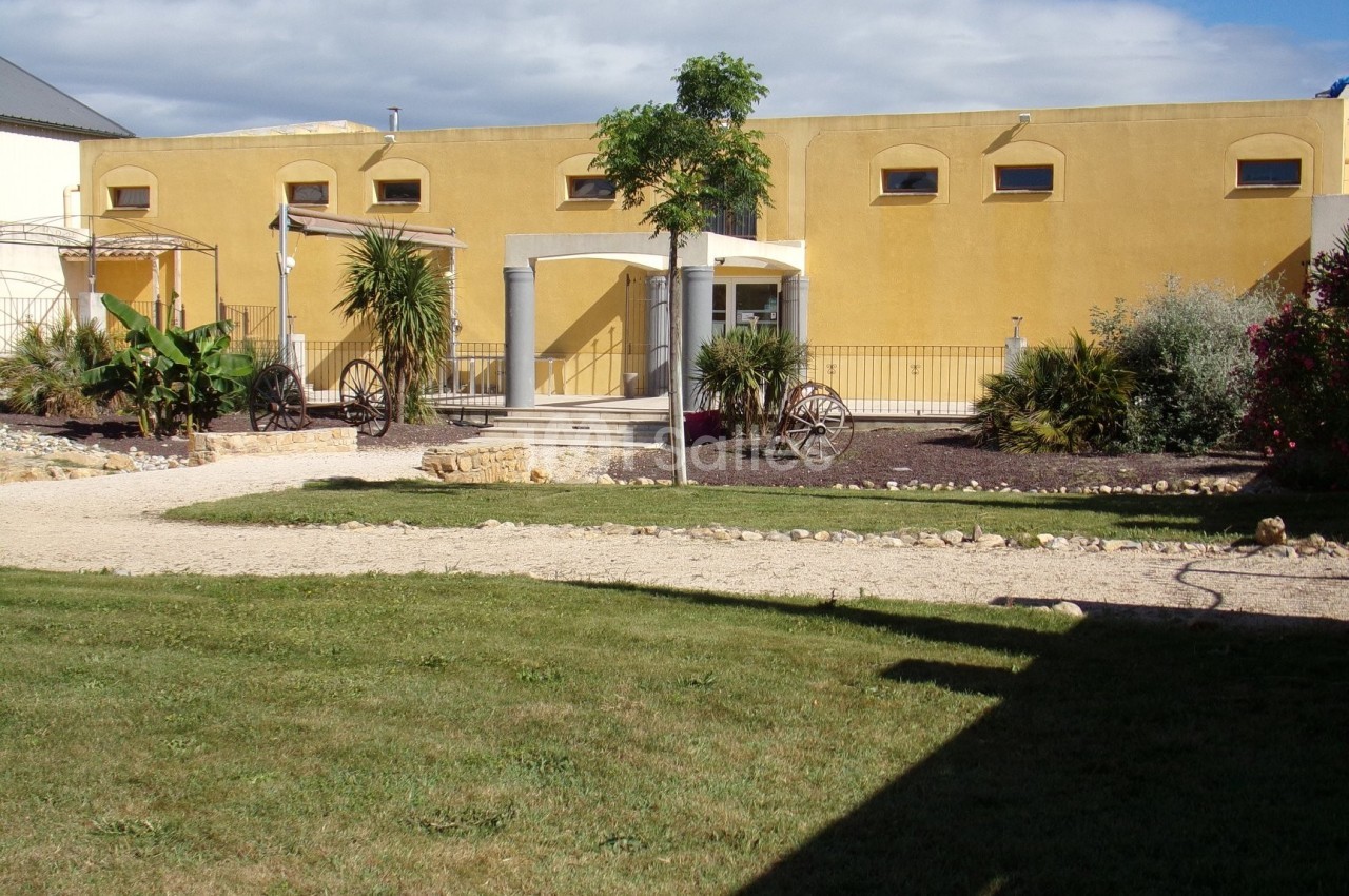 Bâtiment jaune avec des arches, entouré de végétation, d'une allée en gravier et d'une pelouse au premier plan.