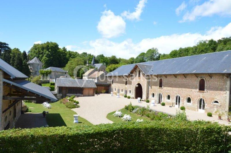 Vue d'un domaine avec bâtiments en pierre, cour gravillonnée, pelouses et arbres sous un ciel bleu.