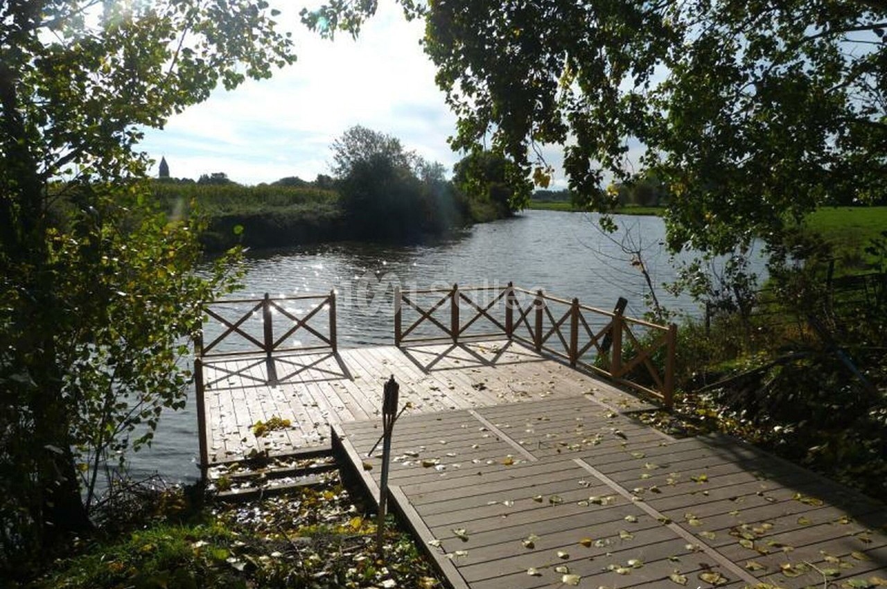 Ponton en bois bordé de barrières, donnant sur une rivière entourée de verdure et d'arbres sous une lumière naturelle.