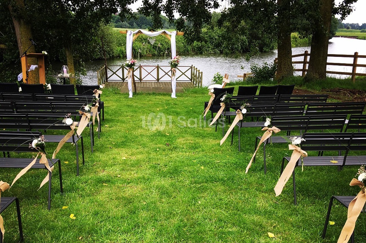 Chaises alignées en extérieur face à une arche décorée, disposées pour une cérémonie près d'un plan d'eau entouré de verdure.