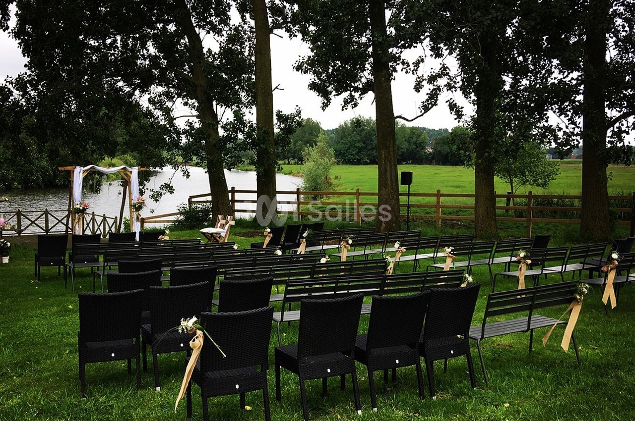 Chaises noires disposées en rangées sur une pelouse, face à une arche décorée près d'un étang entouré d'arbres.
