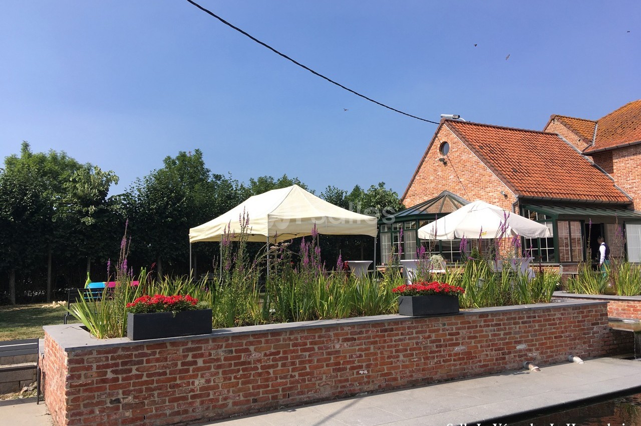 Terrasse ensoleillée avec parterres de fleurs, parasols blancs et bâtiment en briques rouges à l'arrière-plan.