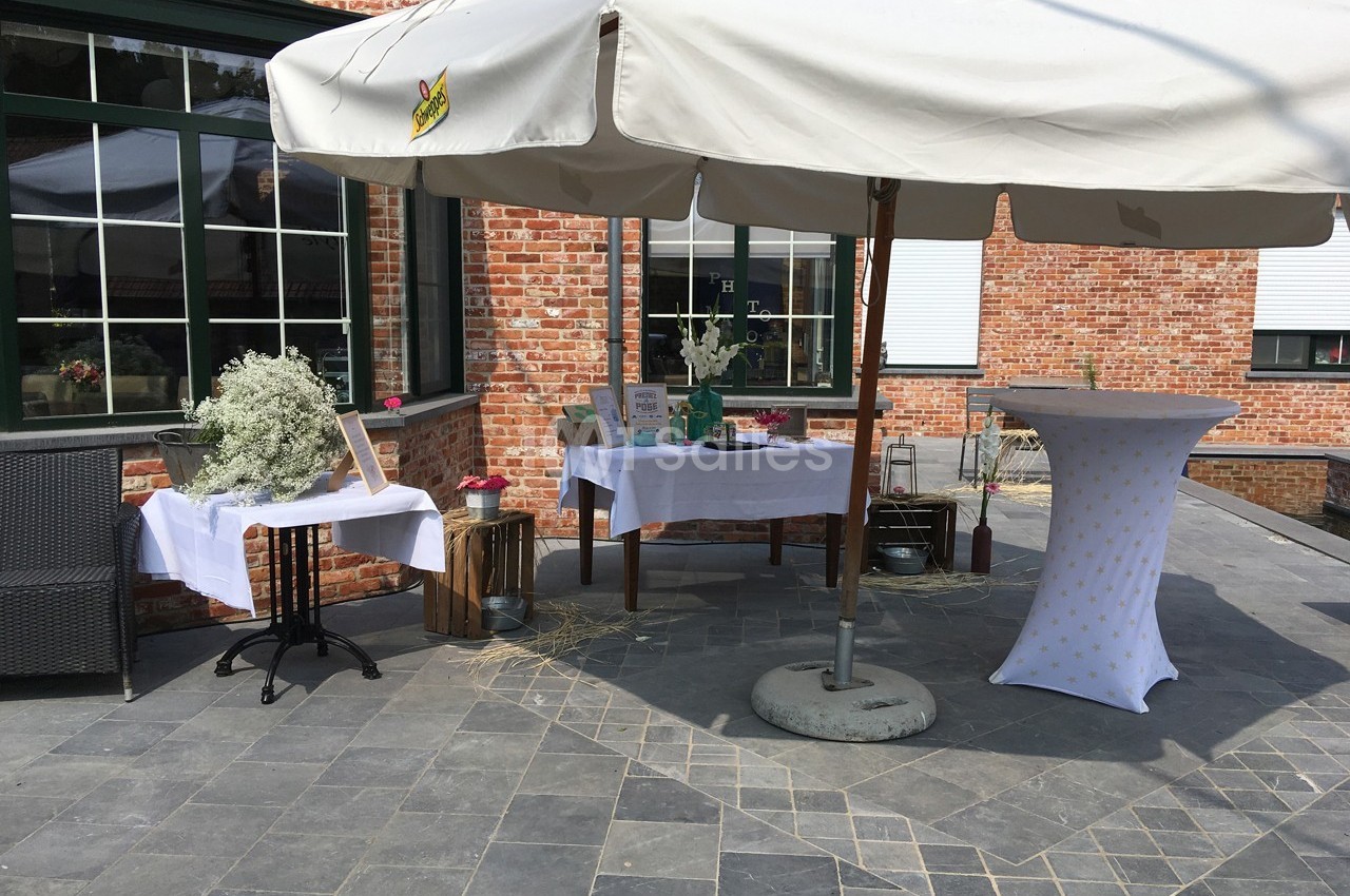 Terrasse avec tables décorées sous un parasol blanc, devant un mur en briques rouges et des fenêtres vitrées.