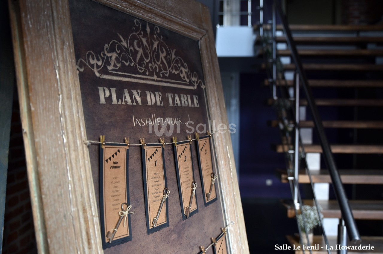 Plan de table affiché sur un panneau en bois avec des cartes suspendues, dans un espace intérieur avec escalier en arrière…