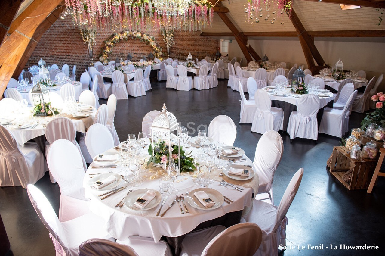 Salle de réception décorée pour un mariage, avec tables rondes dressées, chaises blanches et guirlandes florales suspendues.