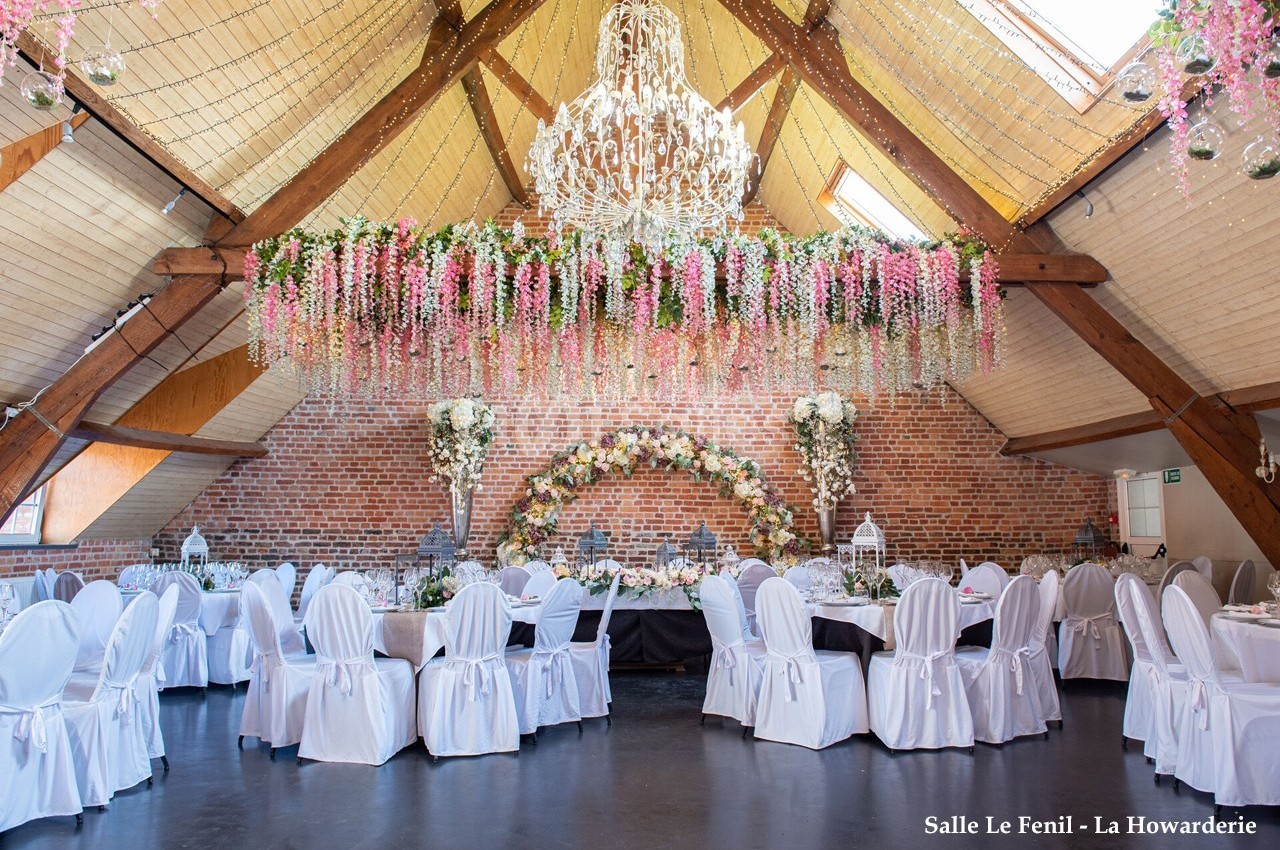 Salle de réception avec tables dressées, chaises blanches, arche florale et lustre sous un plafond en bois.