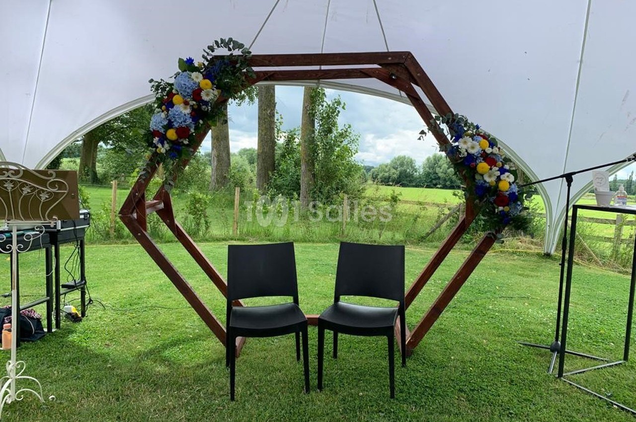 Deux chaises noires devant une arche hexagonale décorée de fleurs colorées, installées sur une pelouse sous une tente…