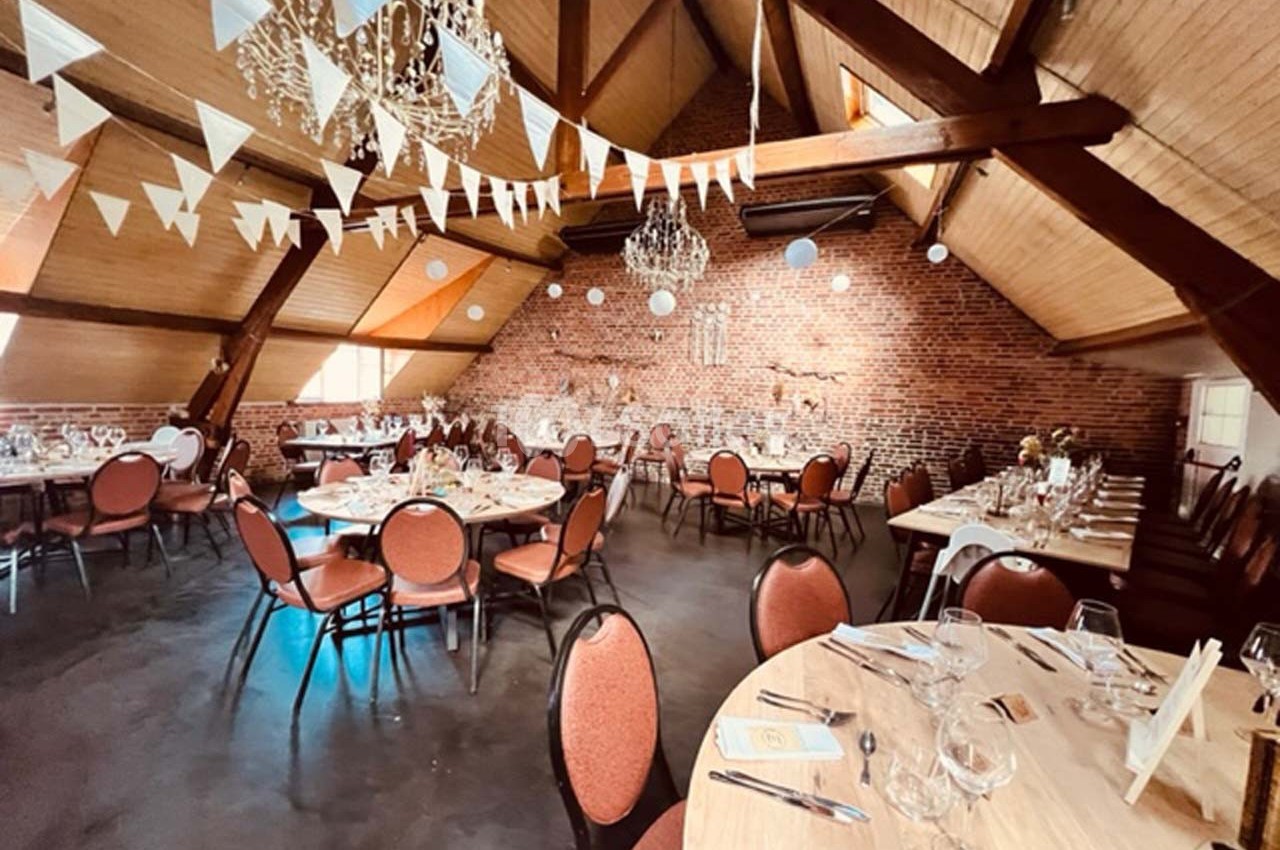 Salle de réception avec charpente apparente, tables dressées, guirlandes de fanions et décorations lumineuses suspendues.