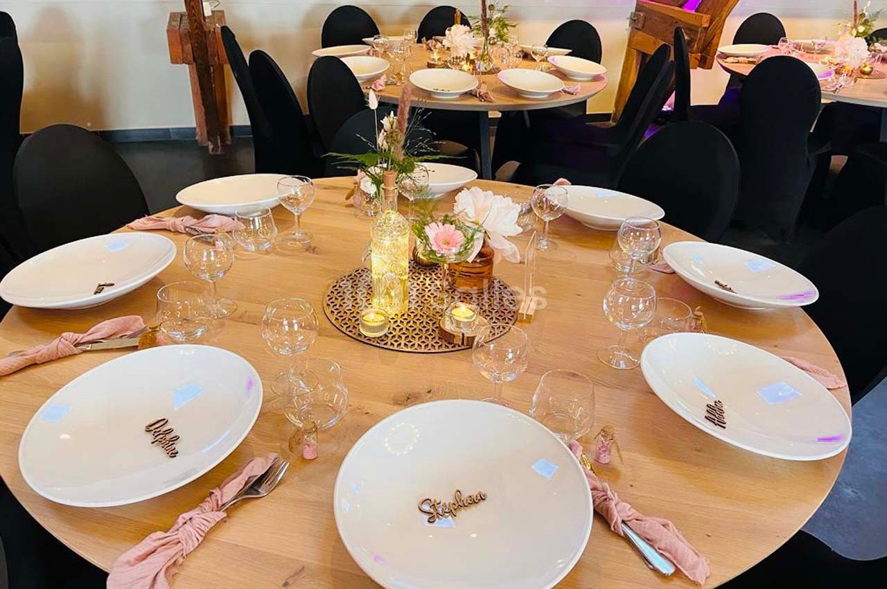 Table ronde en bois décorée pour un événement, avec assiettes, serviettes roses, bougies et fleurs au centre.