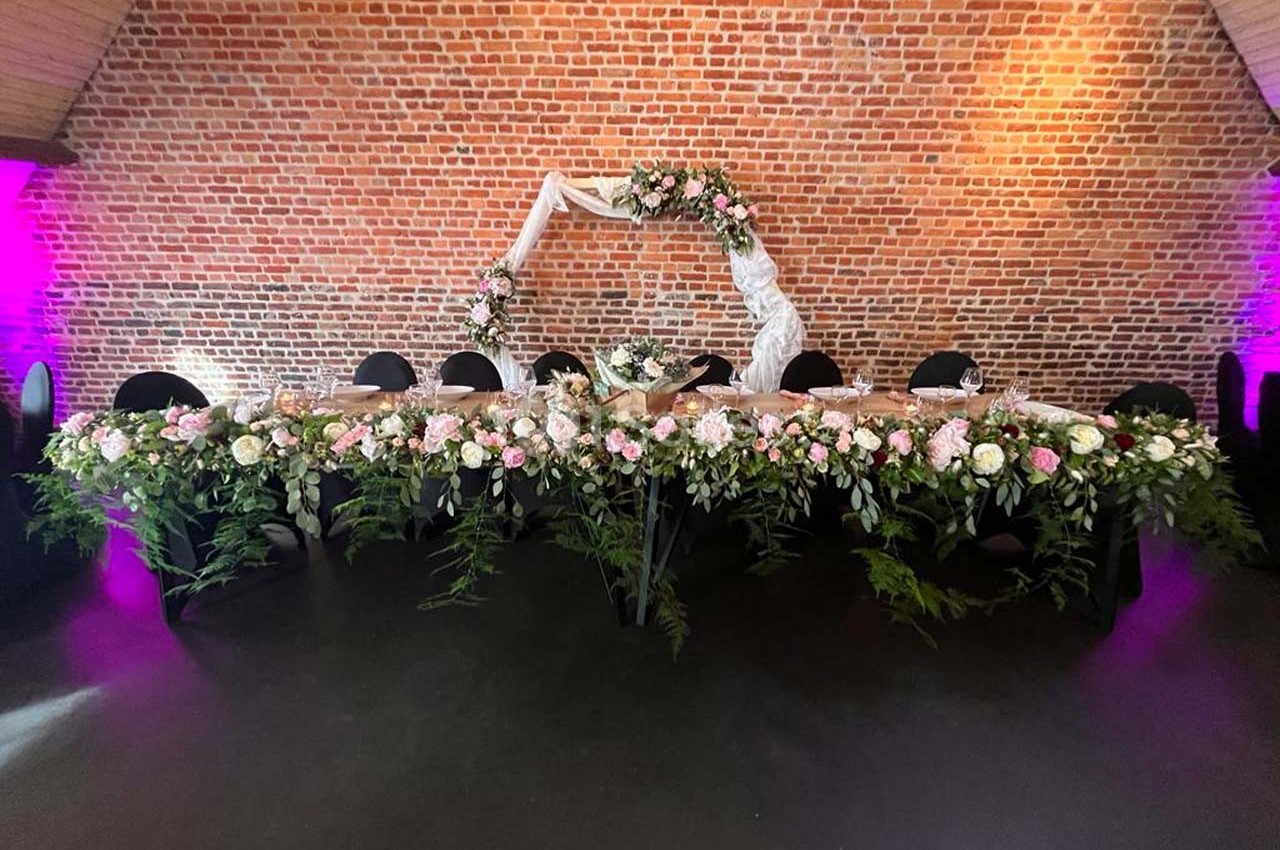 Table décorée pour un événement avec des fleurs roses et blanches, devant un mur de briques éclairé en lumière violette.