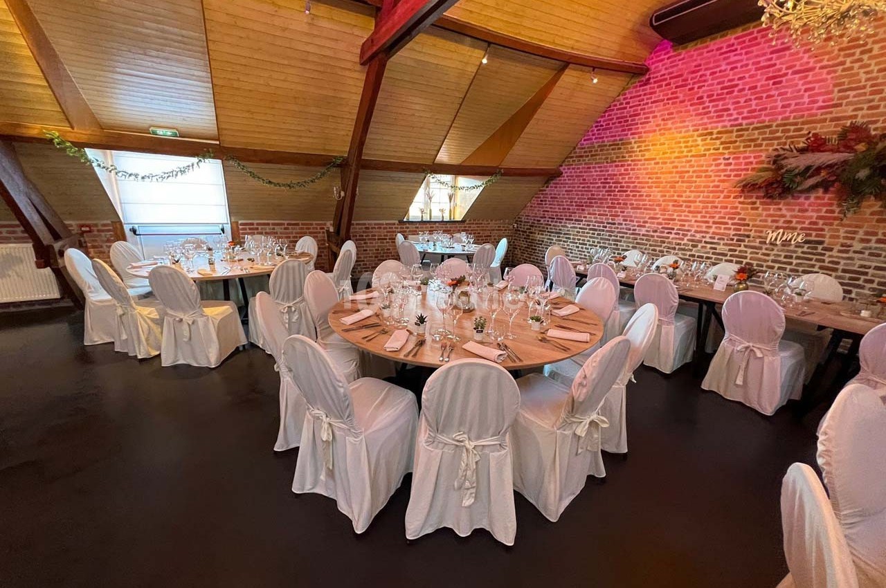 Salle de réception avec tables rondes dressées, nappes blanches et décoration murale en briques et feuillage.